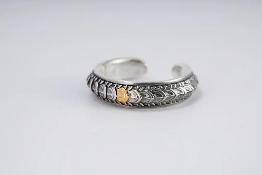 Anillo plateado labrado con un pequeño detalle dorado sobre fondo gris claro
